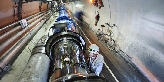Person mit Helm, die sich im Tunnel des Large Hadron Colliders befindet und einen Teil der Beschleunigerstrecke anguckt. Im Hintergrund ist ein Fahrrad zu sehen.