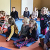 A group of children sits on mats on the floor and looks tensely in one direction. Adults sit in the background.