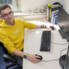 A young man in a yellow sweater sits at a desk with a computer and holds the mouse with his right hand