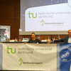 Three men hold a TU Dortmund banner in their hands. The chancellor also holds a prize in his hand.