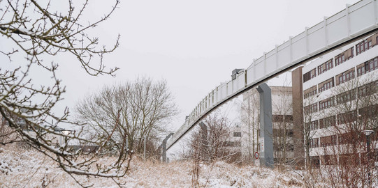 Verschneiter Nordcampus mit einem Gebäude, der H-Bahn-Strecke und Bäumen.