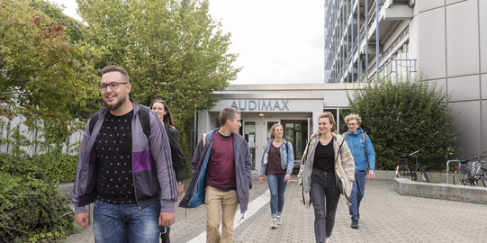 Sechs Studierende laufen teilweise zueinander gewandt aus dem Audimaxgebäude heraus.