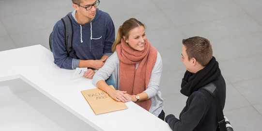 Drei Personen stehen neben einenander an einer Theke. Die Person in der Mitte lacht die Person rechts an und hat ihre Hände auf einen großen Umschlag liegen. Die Person links hat einen kleinen Zettel mit einer Wartenummer in der Hand.