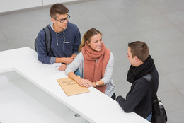 Drei Personen stehen neben einenander an einer Theke. Die Person in der Mitte lacht die Person rechts an und hat ihre Hände auf einen großen Umschlag liegen. Die Person links hat einen kleinen Zettel mit einer Wartenummer in der Hand.