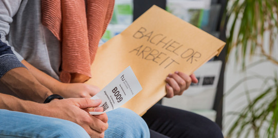 Close-up of two people sitting nect to each other. One is holding an envelope, the other a small piece of paper with a number printed on it
