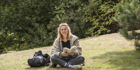 A female student sits cross-legged on a meadow with a book in her hand.