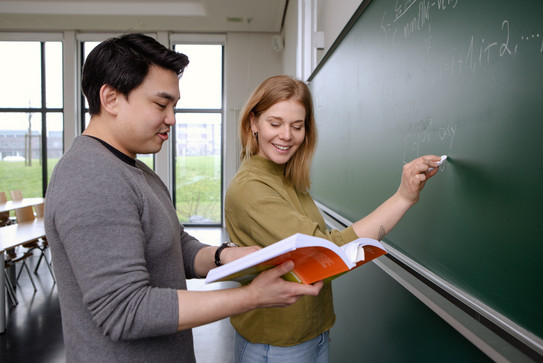 Zwei Studierende an der Tafel, er hält Buch, sie schaut rein, beide lächeln