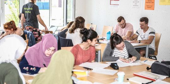 Mehrere Personen sitzen an Tischen und haben Unterlagen vor sich. Eine Person steht und trägt ein T-Shirt mit der Aufschrift "HelpDesk-Mathe".